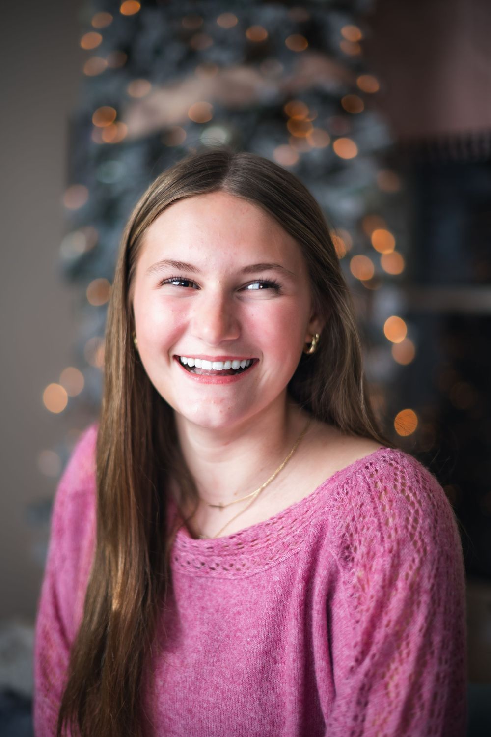 A person in a pink sweater smiles warmly against a blurred background with twinkling bokeh lights.