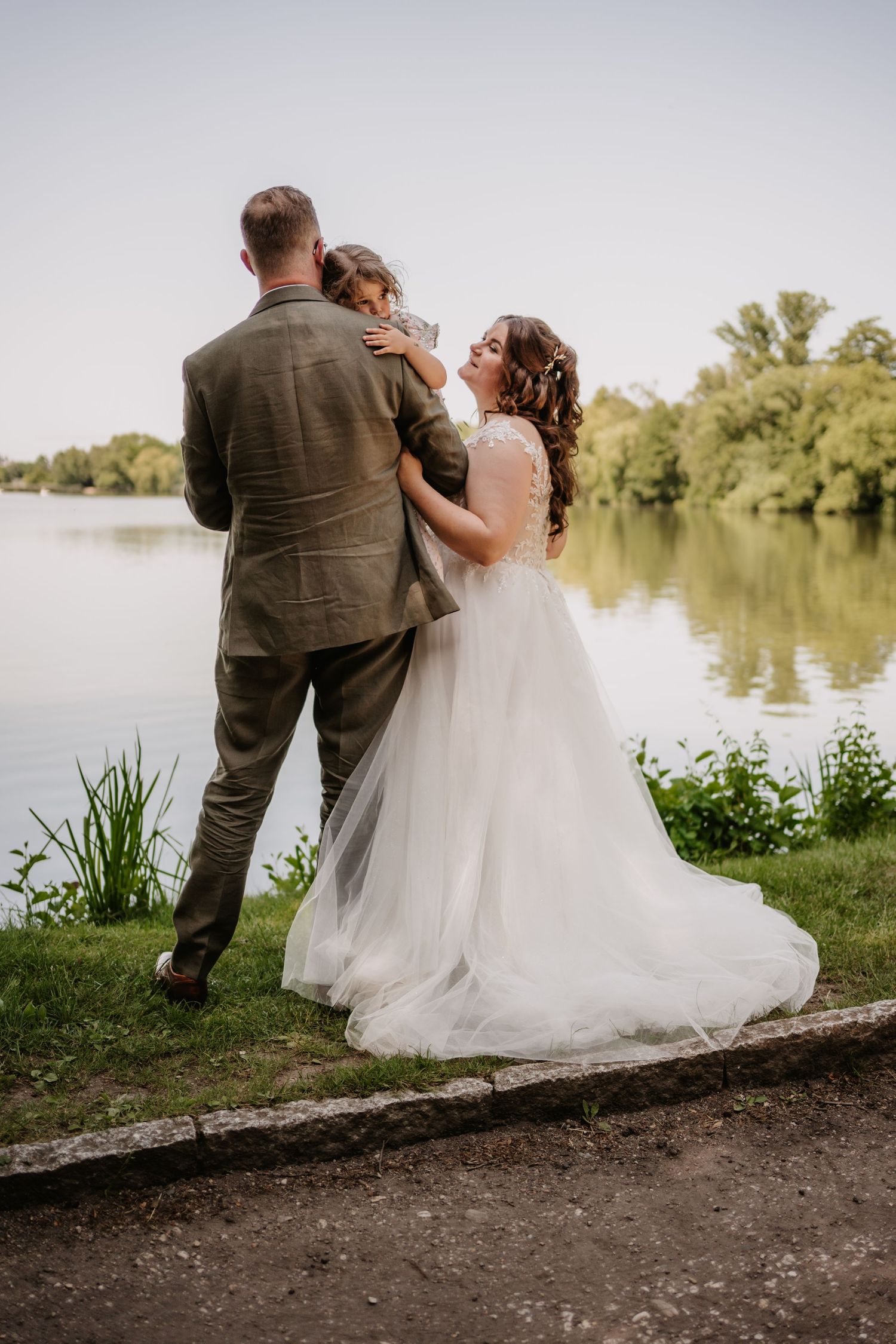 Romantic couple in wedding attire sits together on a log by a serene lake during sunset.