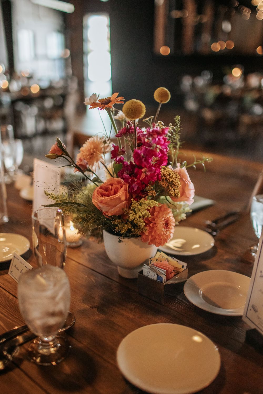 Elegant table setting with pink and peach floral centerpieces in white vases on a rustic wooden table with candlelight.
