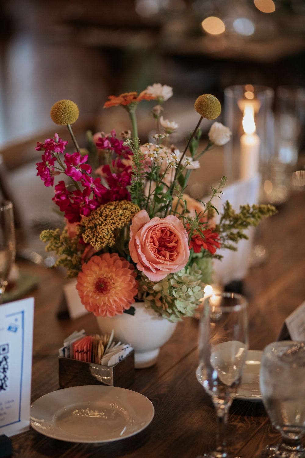 Close-up of vibrant coral and pink floral arrangements with roses and dahlias on rustic wooden dining tables.