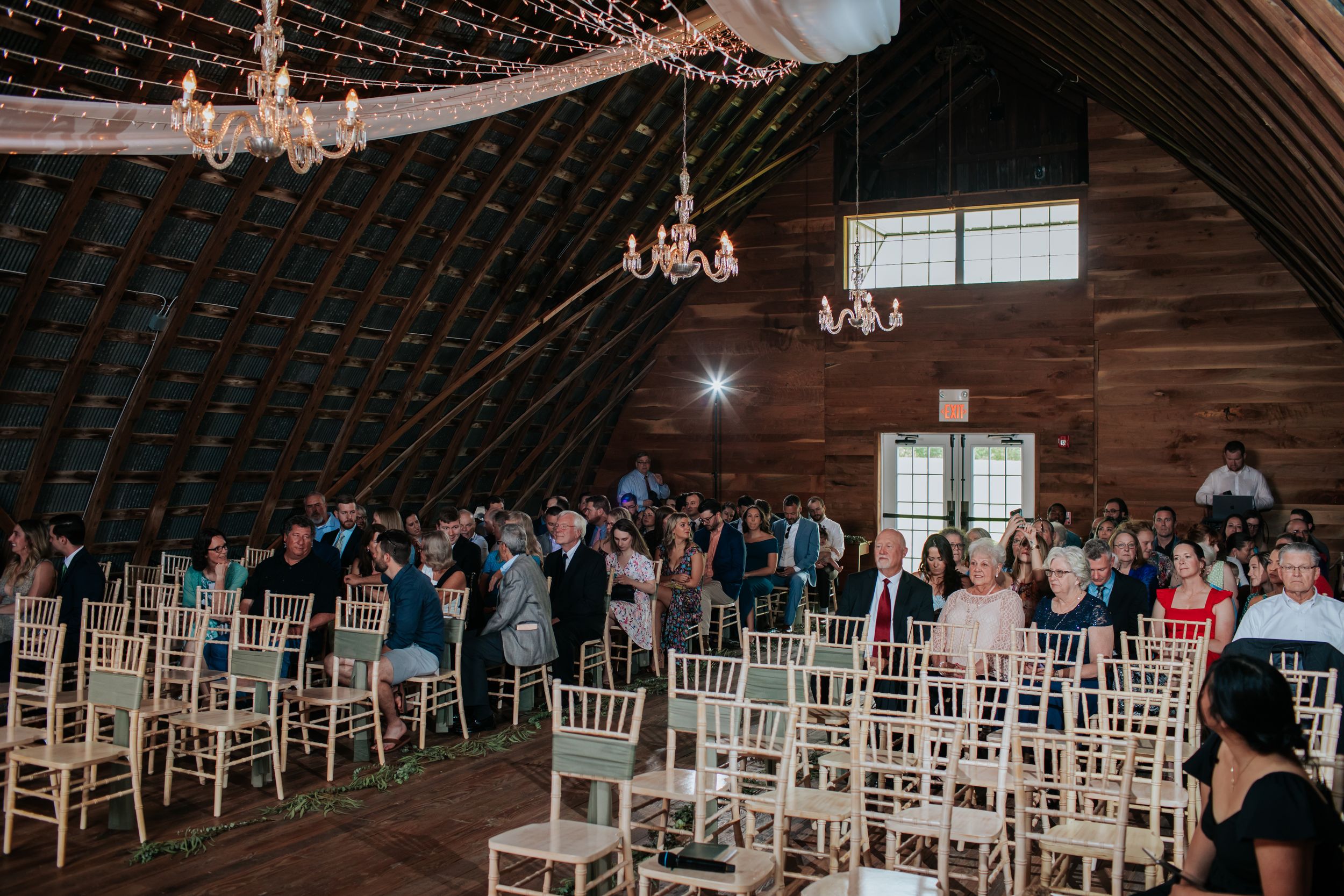 Wedding ceremony setup in rustic barn with white chairs and hanging string lights.