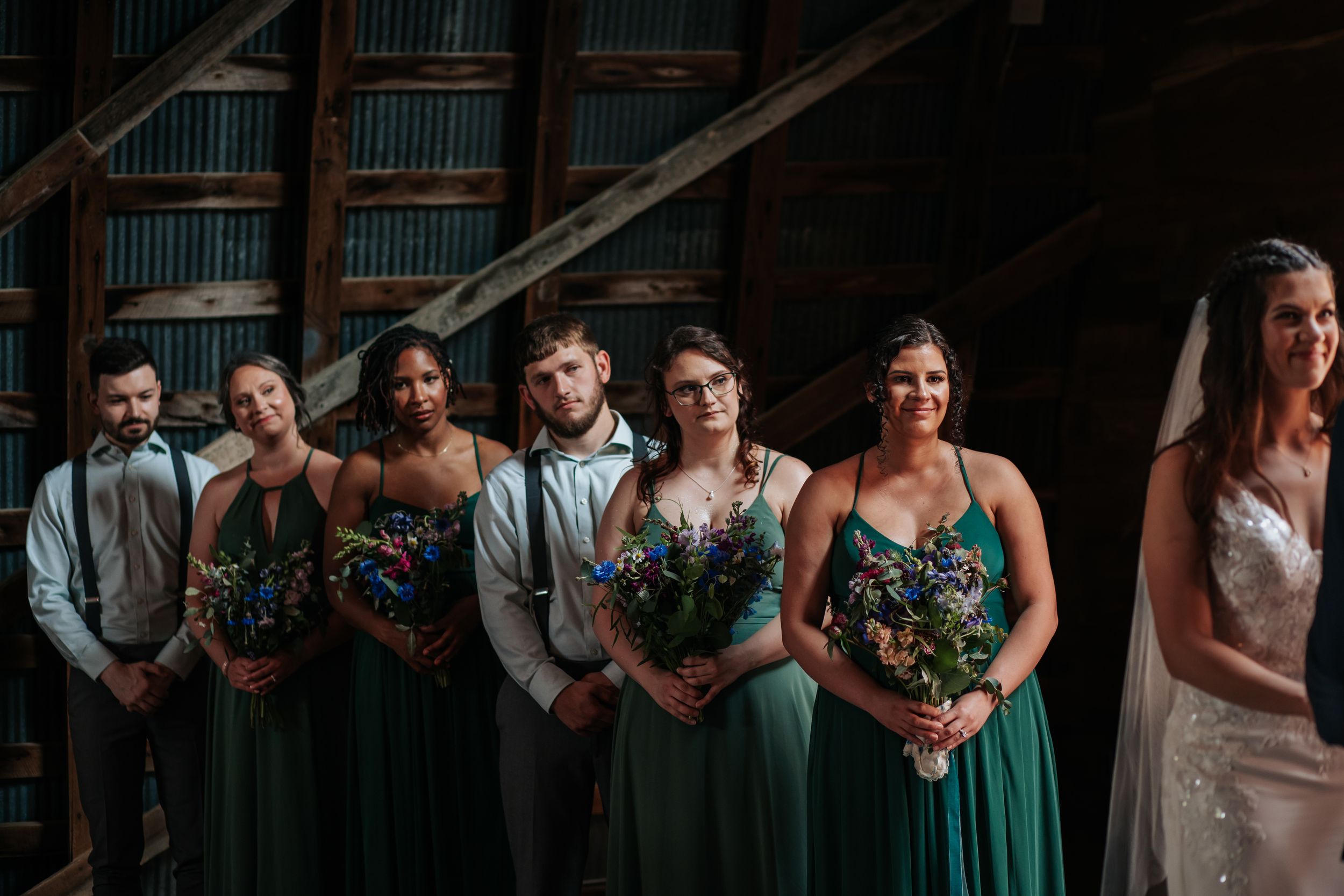 Wedding party lineup with bridesmaids in emerald green dresses and groomsmen in dark suits against rustic backdrop.