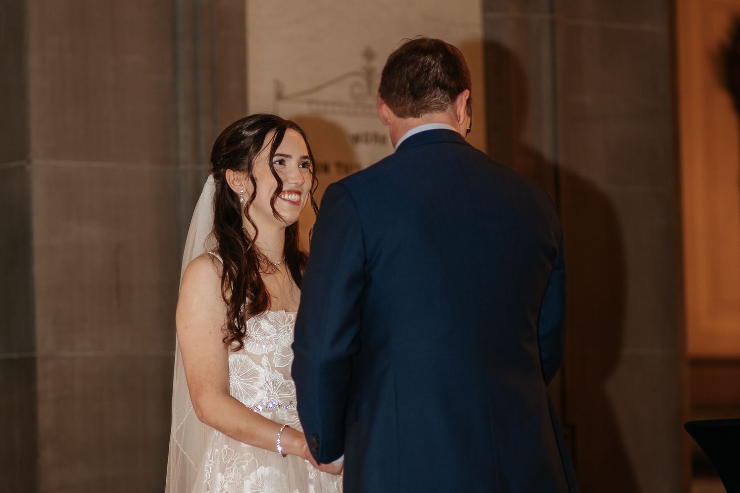 A bride and groom share their first dance in an elegant ballroom with warm lighting and stone architecture.