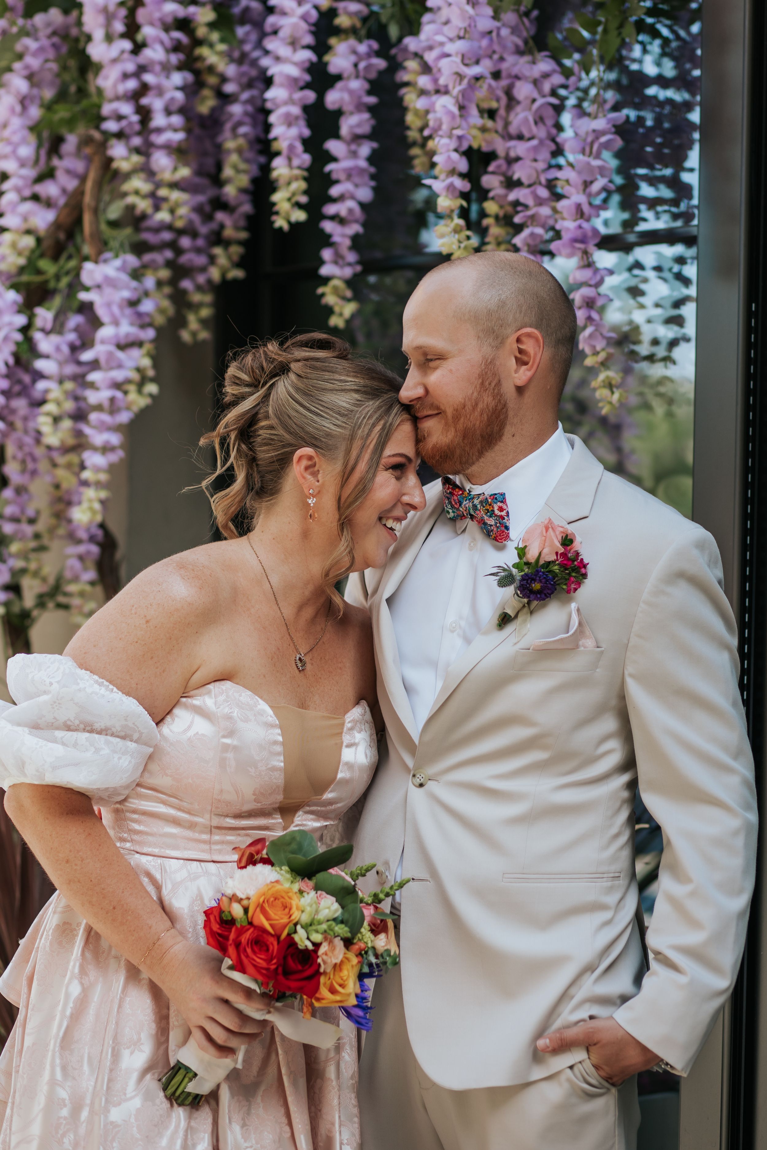 A romantic wedding couple shares tender moments under hanging purple wisteria flowers in a garden setting.