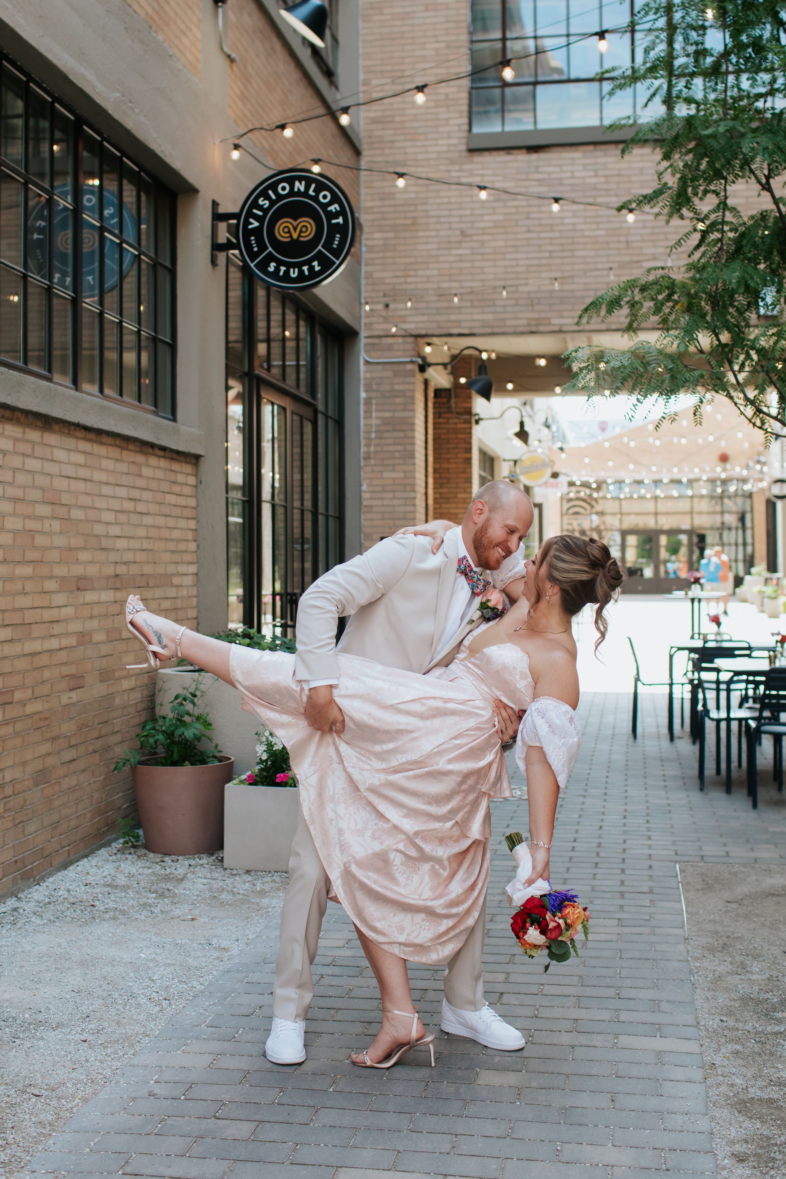 A couple in wedding attire performs playful dance poses together on a brick-lined walkway.