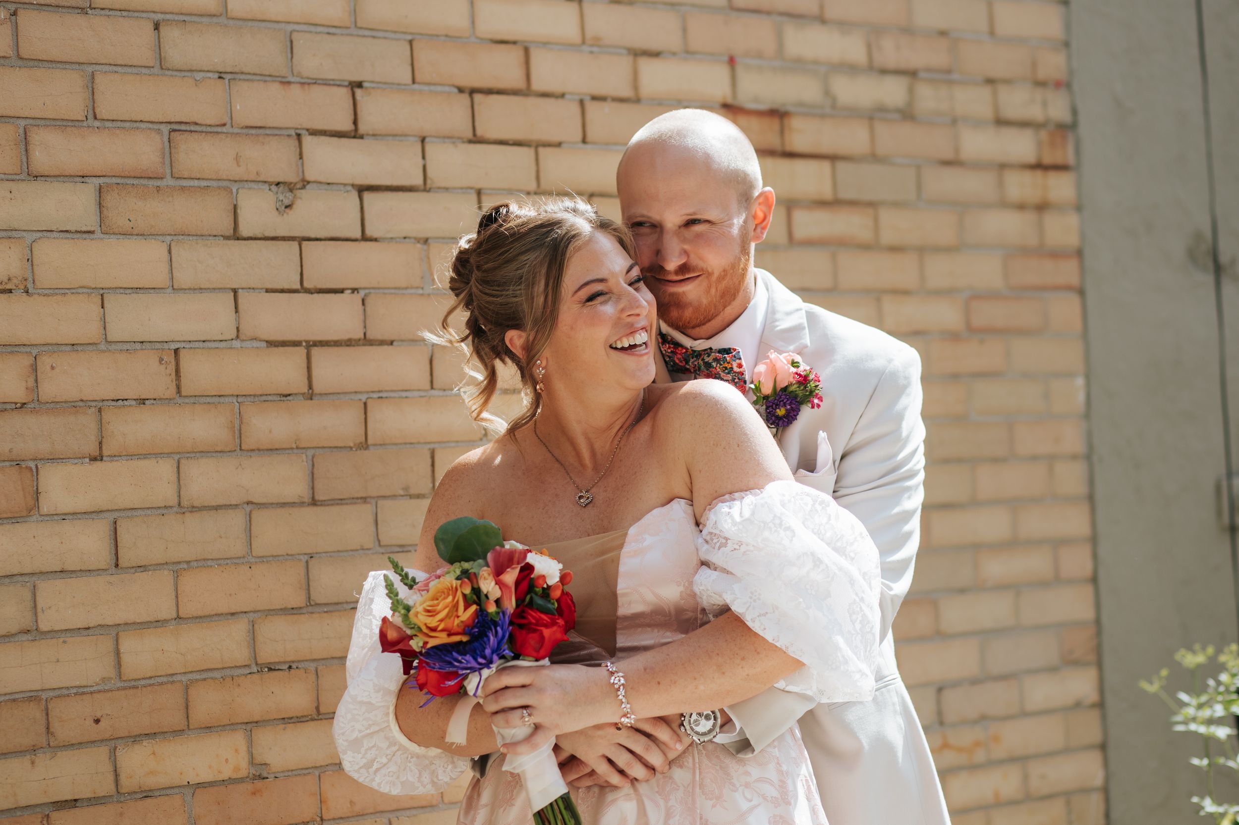 A romantic wedding couple embraces against a brick wall backdrop while holding a vibrant bouquet of flowers.