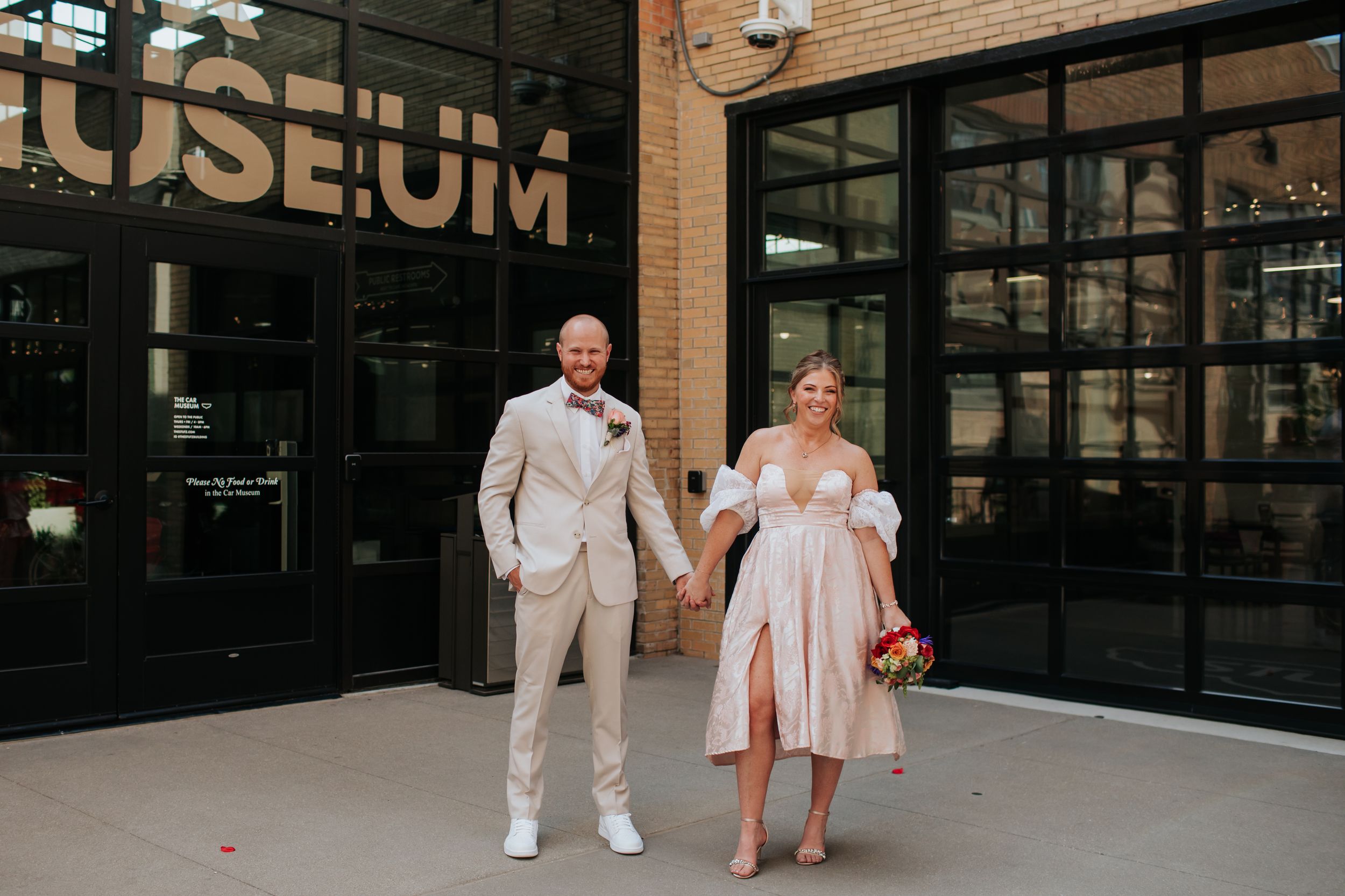 A couple poses together outside a museum entrance wearing light colored formal attire.