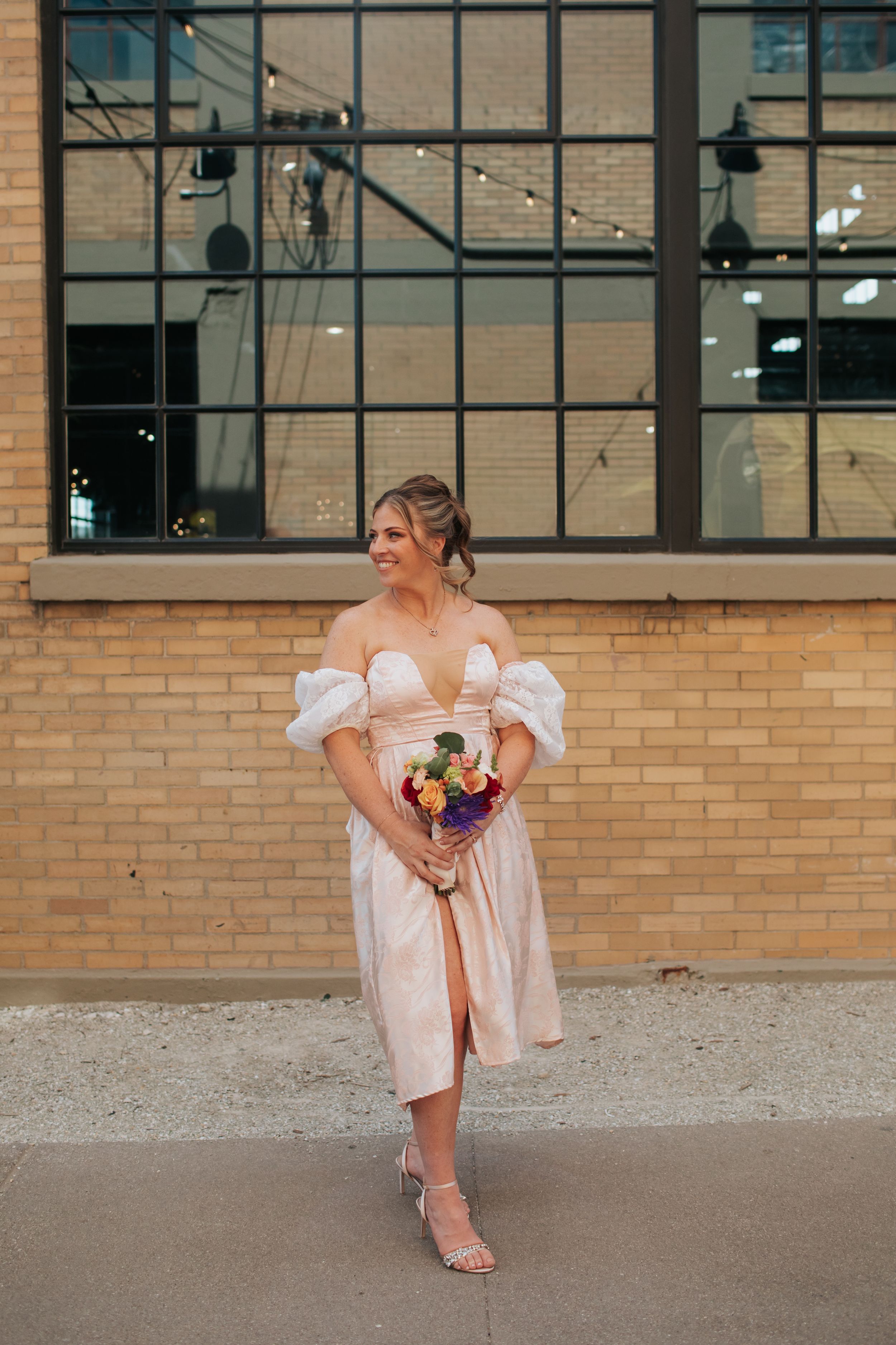 A bride in a white off-shoulder dress poses playfully against a brick wall with industrial windows.
