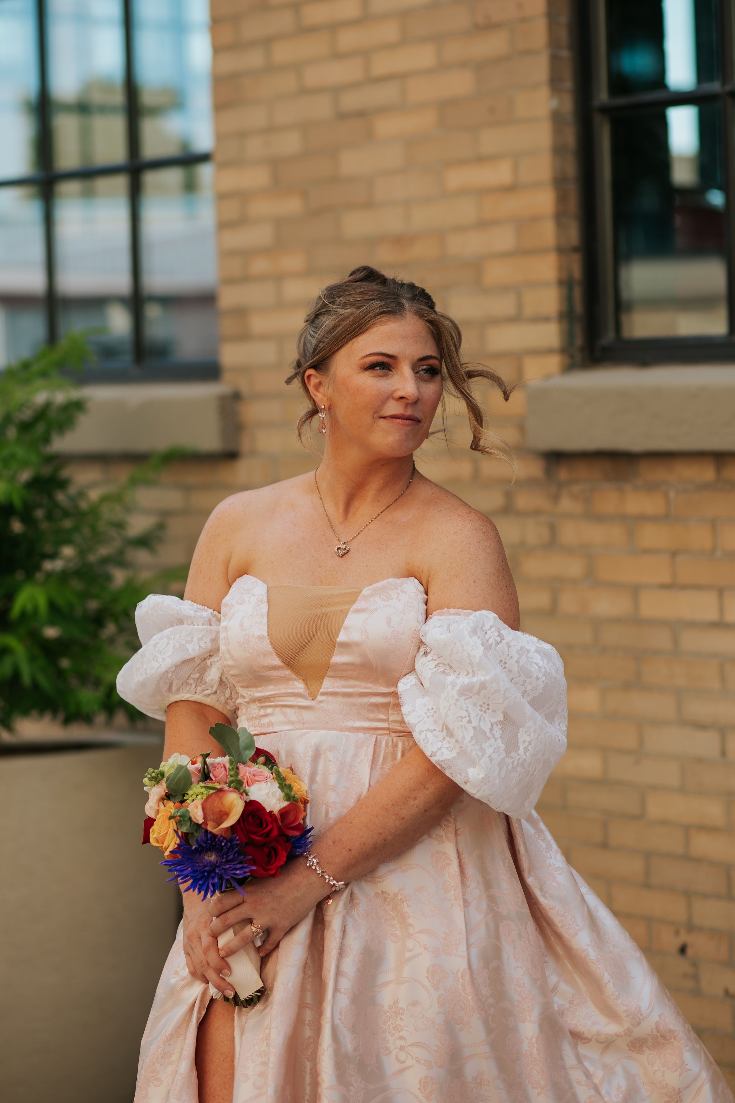 Bride in a romantic blush pink off-shoulder gown holds a vibrant bouquet against a brick wall backdrop.