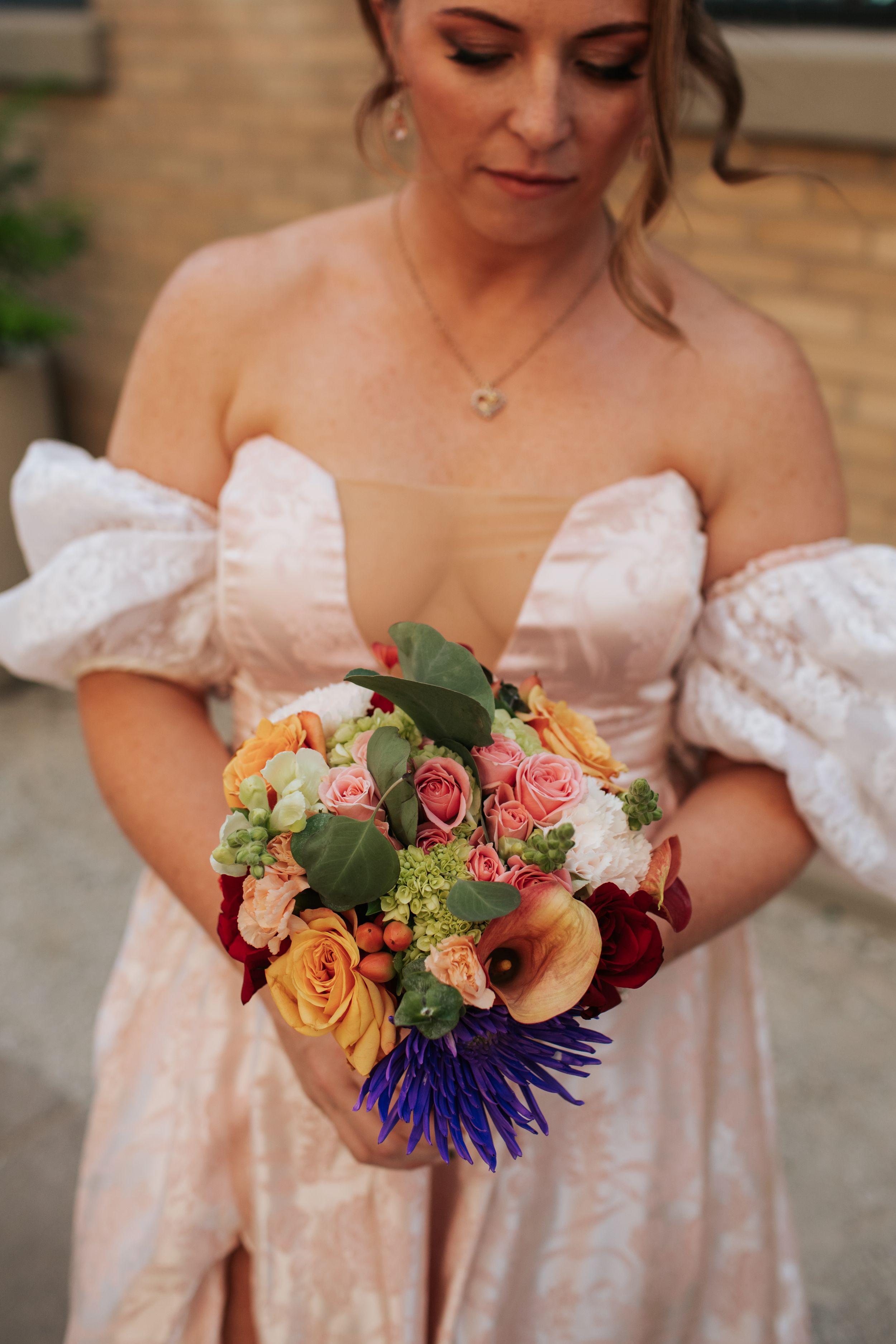 A vibrant wedding bouquet featuring pink roses, orange blooms, and deep purple flowers against a white dress.