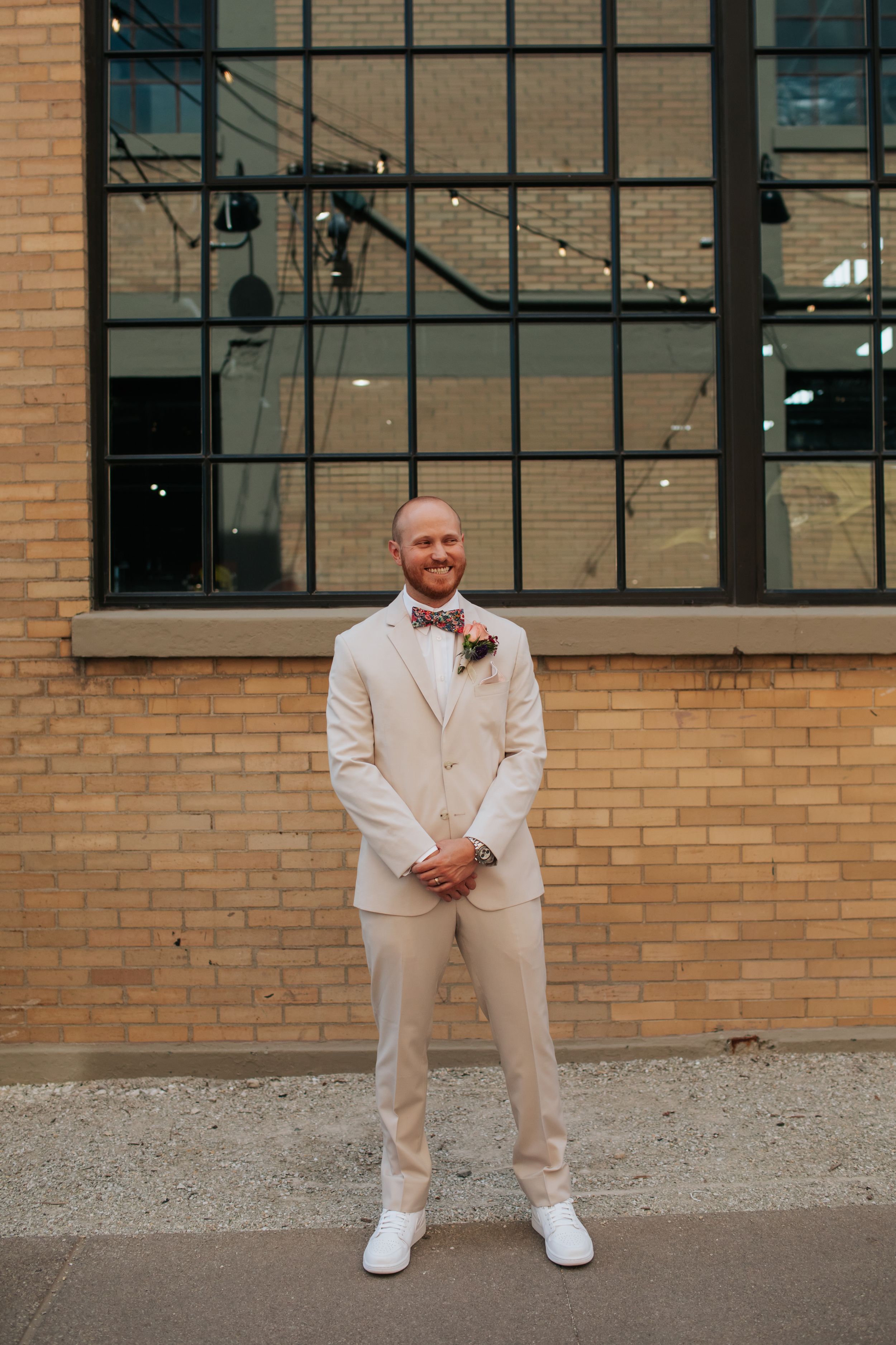 A person in a cream-colored suit poses against a brick building with industrial windows.