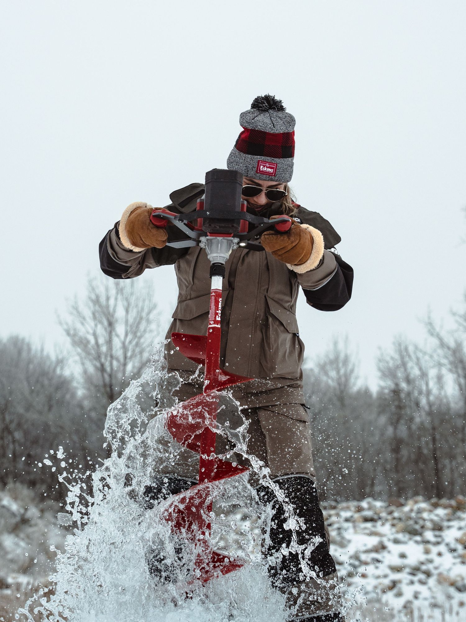 Ice fisherman drilling through frozen lake with red power auger creating dramatic snow spray.