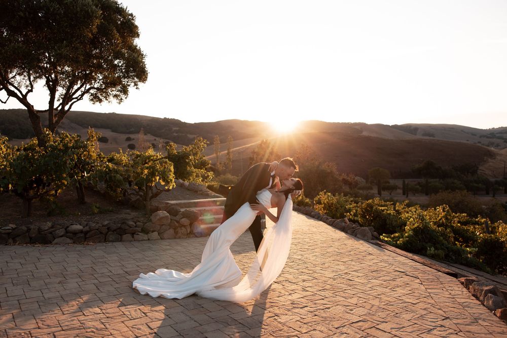 A couple in wedding attire embraces on a stone path at sunset with mountains in the background.