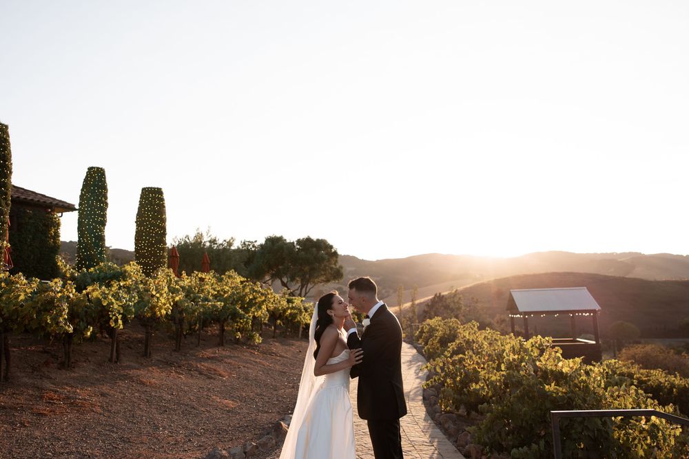 Newlywed couple embraces during golden hour sunset in a scenic vineyard setting with cypress trees in background.