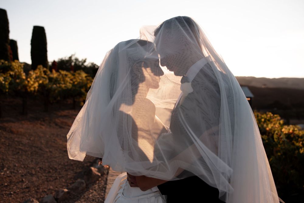 A romantic wedding couple embraces at sunset with a flowing veil creating an ethereal silhouette against the sky.
