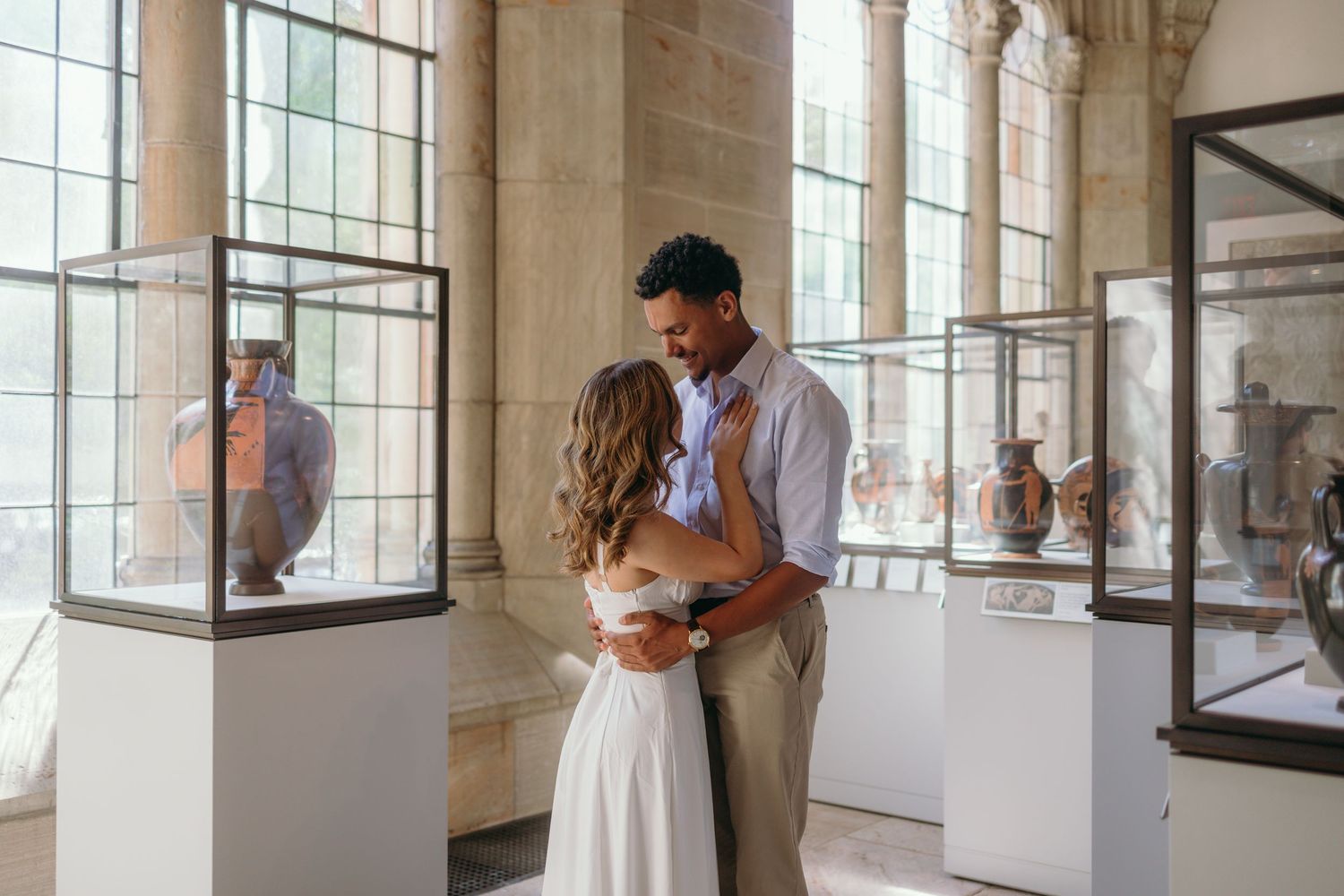 Couple embraces in an elegant museum gallery with glass display cases and tall windows.