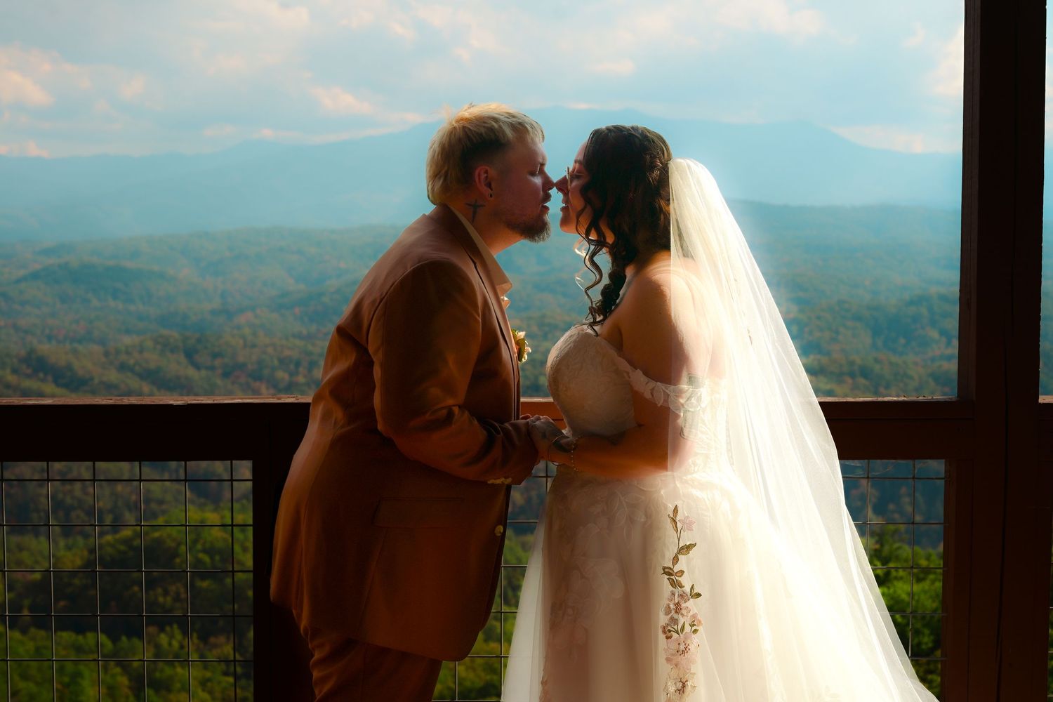 A romantic wedding kiss silhouetted against a scenic mountain vista through large windows.
