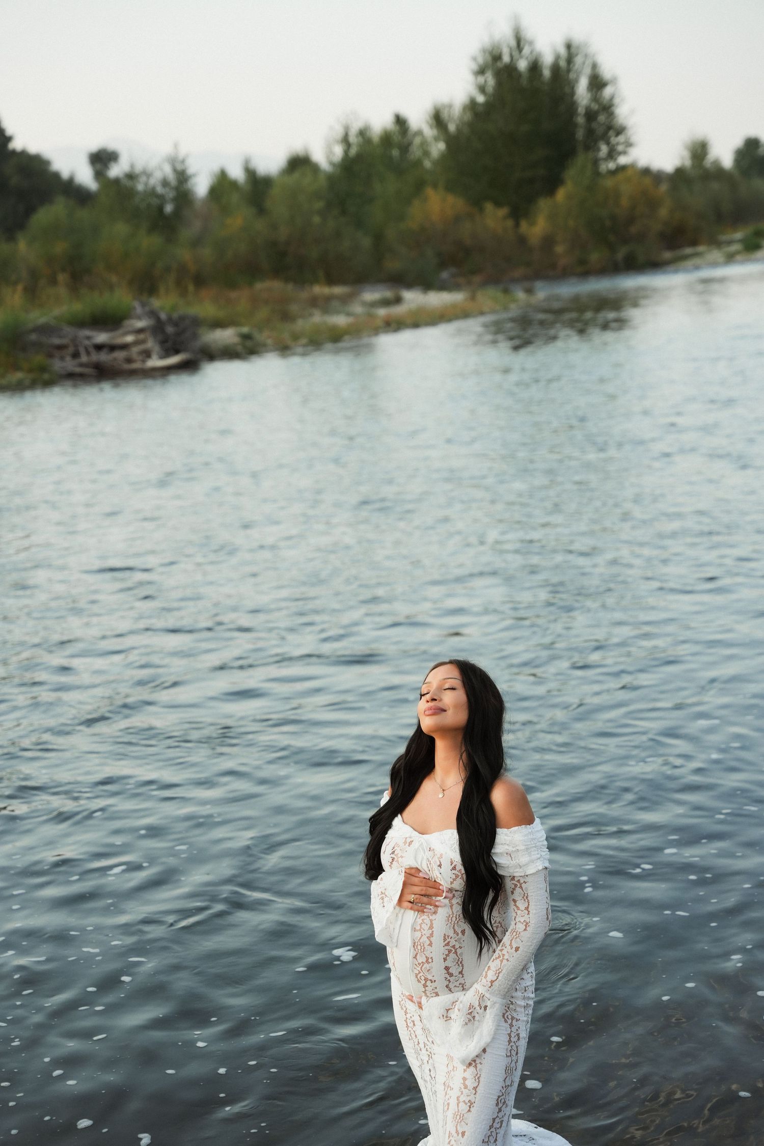 A figure in a white lace dress stands in a lake with dark rippling water and trees along the shoreline.