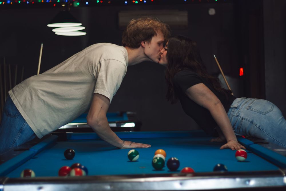 Young couple sharing a kiss leaning over a pool table with billiard balls during dive bar photo session in Yelm WA