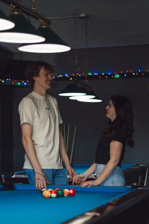Smiling couple holding hands across a blue pool table under pendant lights during romantic photoshoot at Yelm WA dive bar.