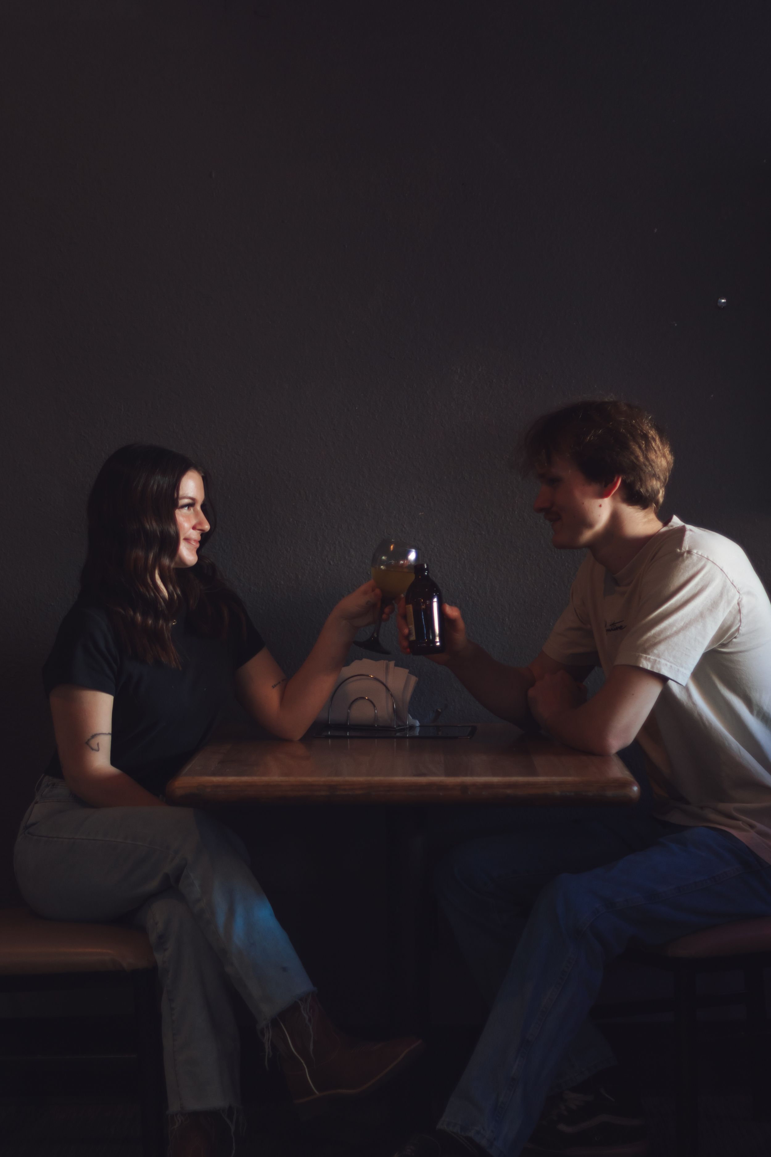 Couple clinking beer bottles together at a dimly lit wooden table, smiling during photoshoot at Tommy's Tavern in Yelm Washington