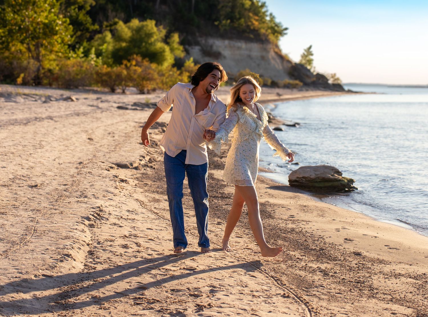 A couple strolls along a sandy beach at sunset with trees and ocean waves in the background.