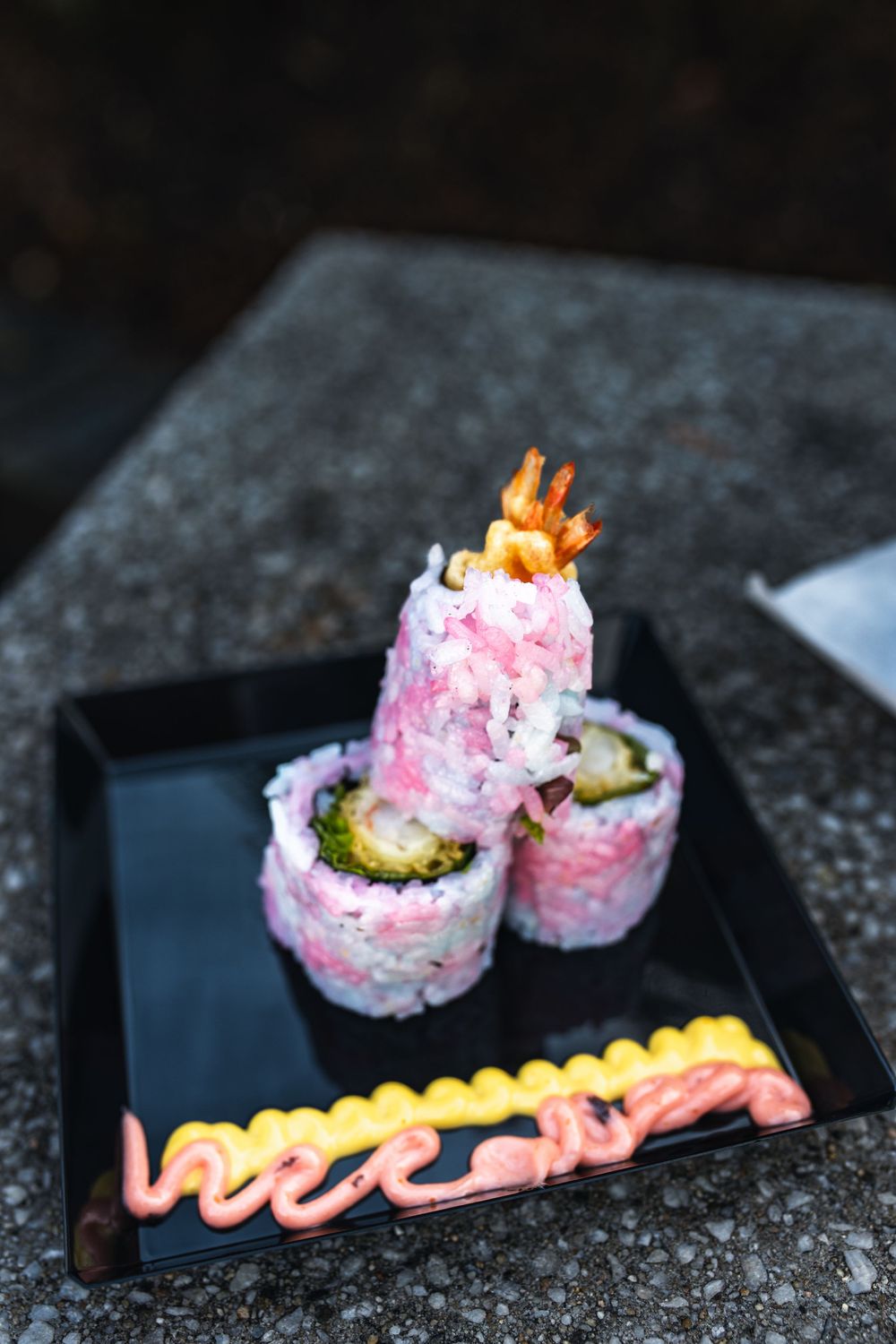 Close-up sequence of pink sushi rolls with tempura shrimp and drizzled sauce on a black plate against dark background.