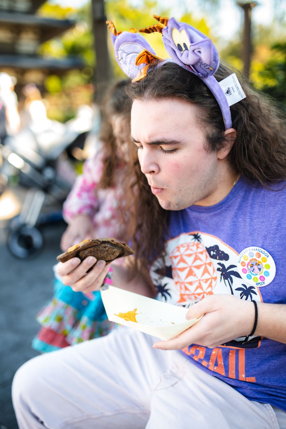A person in a purple bandana and blue t-shirt with tropical designs takes a bite of food outdoors.