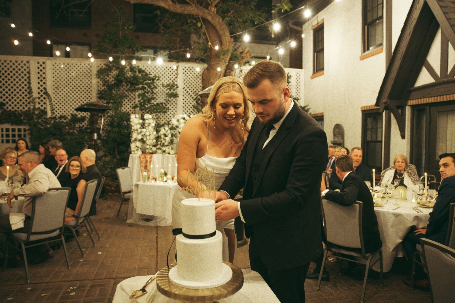 A couple cuts their elegant three-tiered white wedding cake at an outdoor evening reception with string lights overhead.