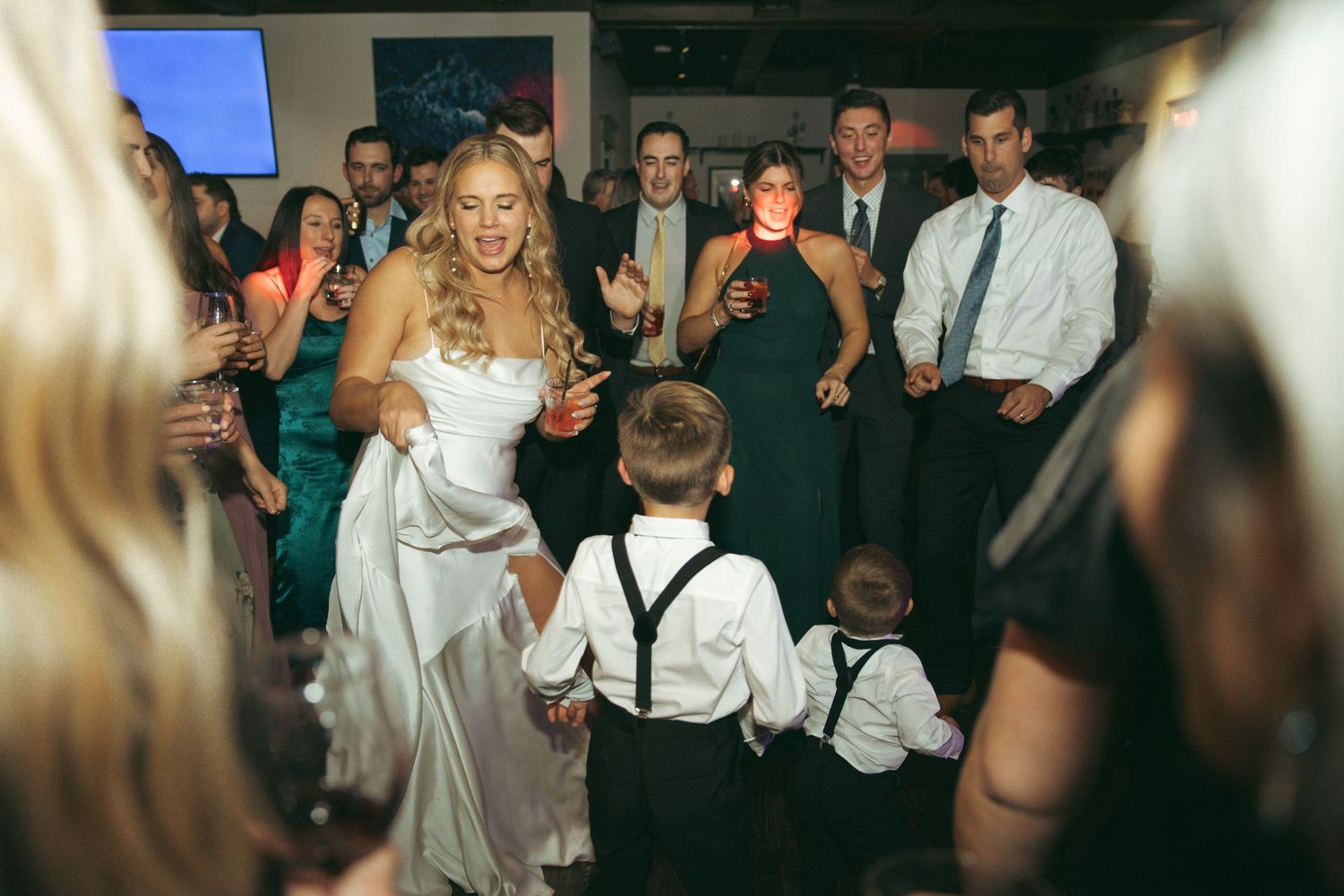 A bride in a white gown dances with two young ring bearers wearing suspenders at a wedding reception.