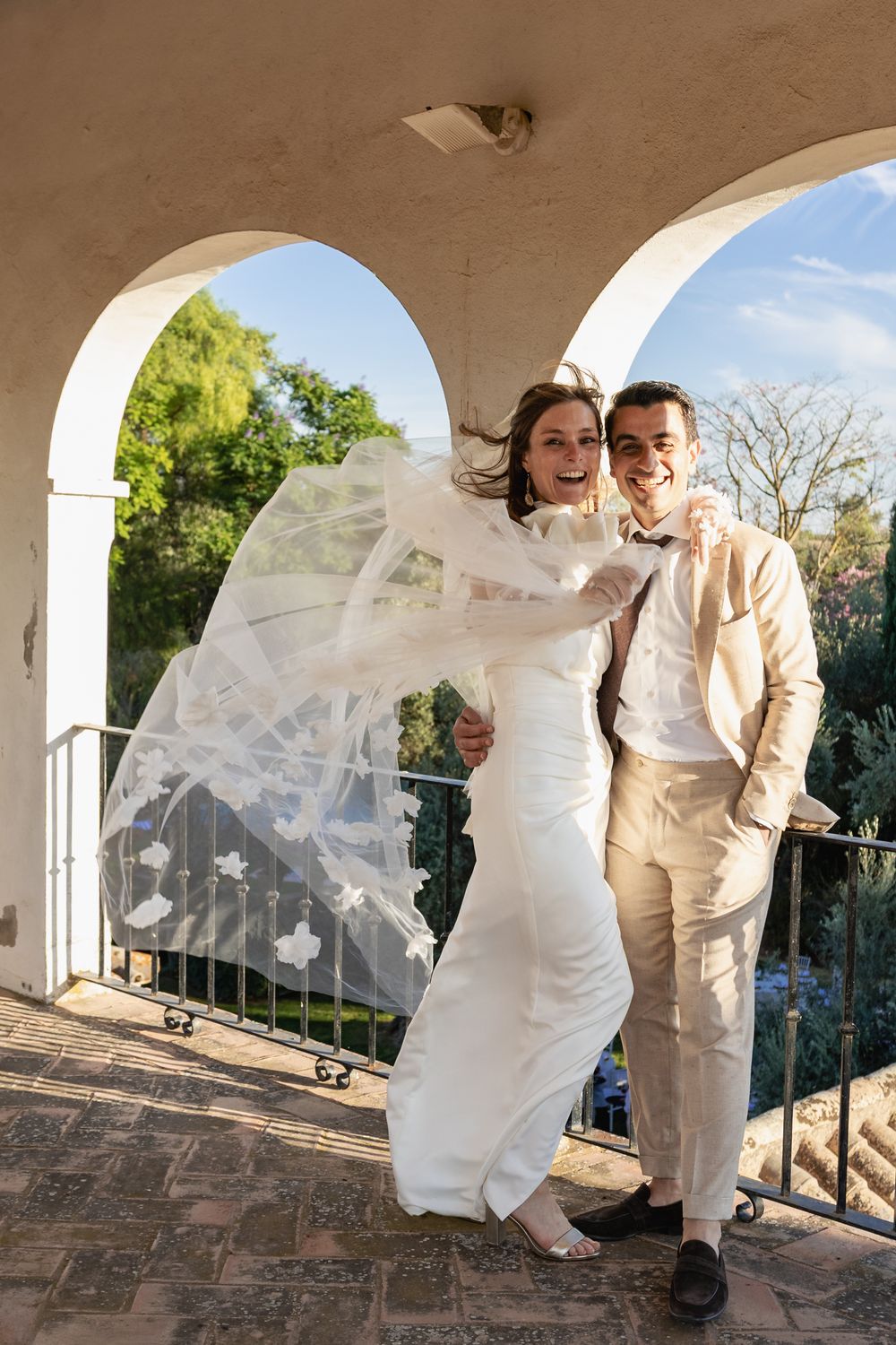 A romantic wedding couple shares playful moments under white stone arches as the bride's veil flows in the breeze.