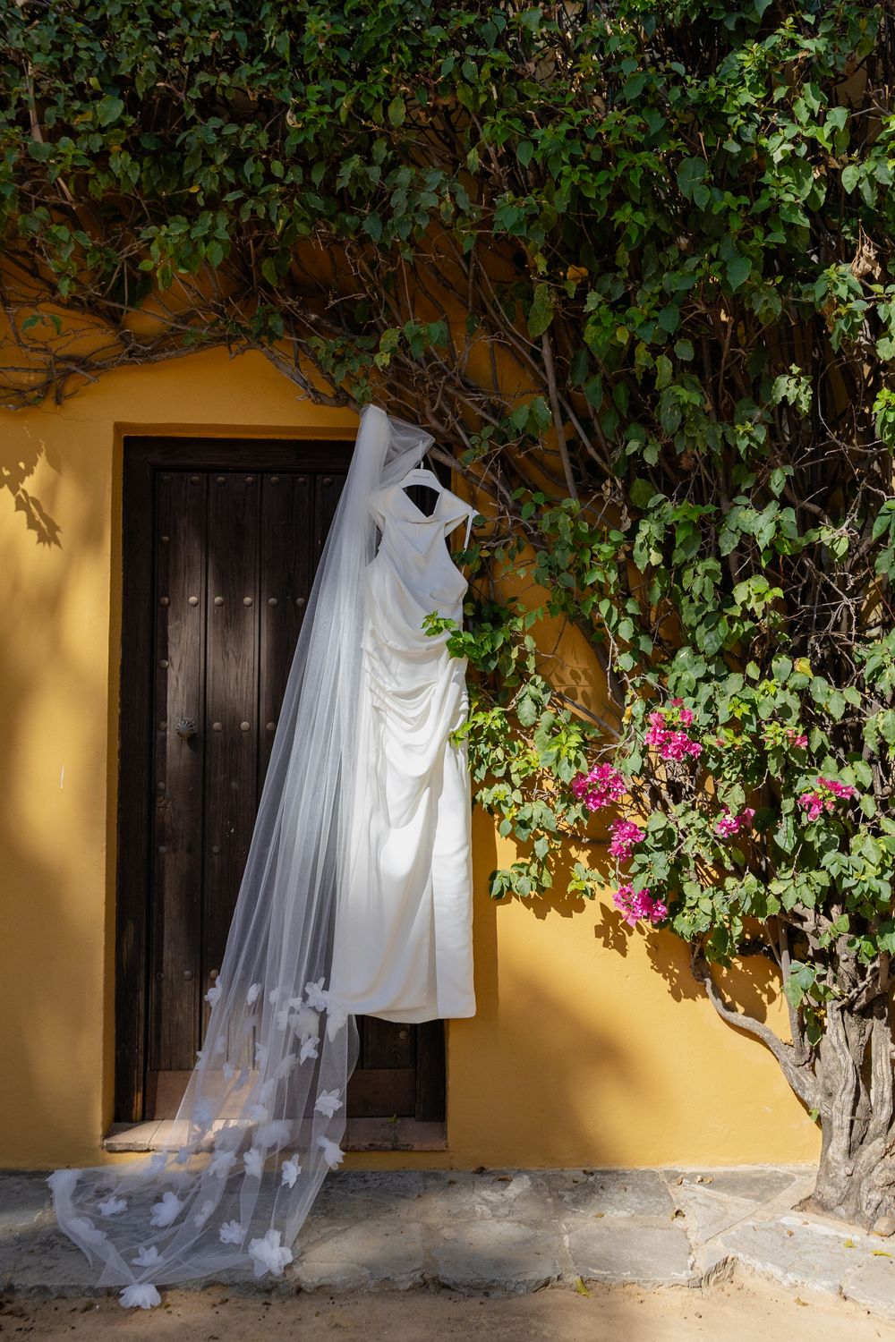 A white wedding dress hangs against a yellow wall with climbing vines and pink flowers in an outdoor setting.