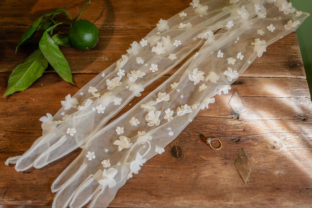 White lace bridal gloves with delicate floral appliques displayed on a rustic wooden surface near green citrus leaves.