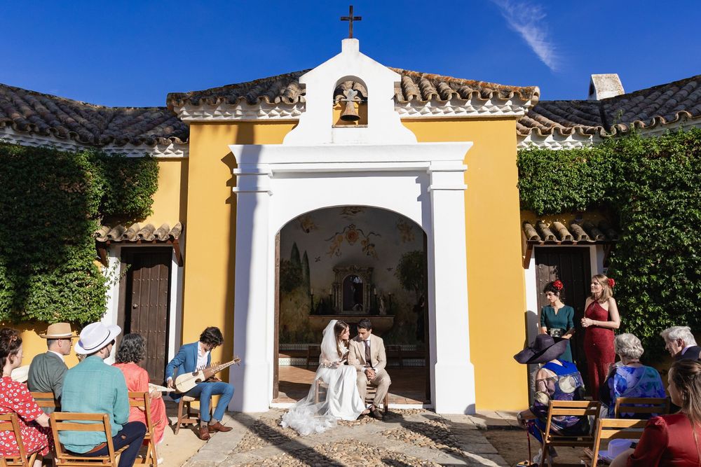 Small outdoor chapel with yellow walls and white archway entrance where people gather for a ceremony in bright sunlight.
