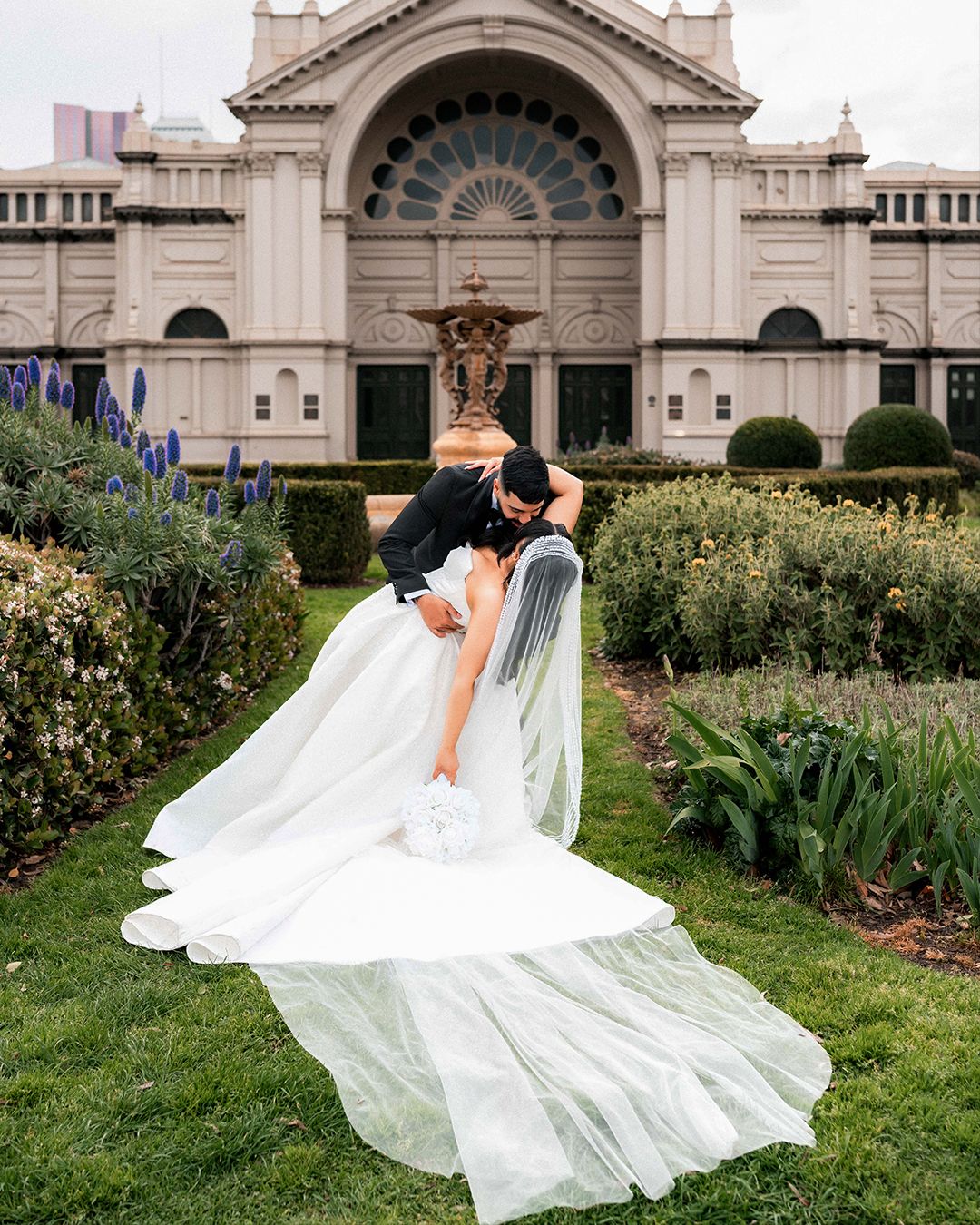 A flowing white wedding veil billows dramatically in the wind against ornate classical architecture and garden landscaping.