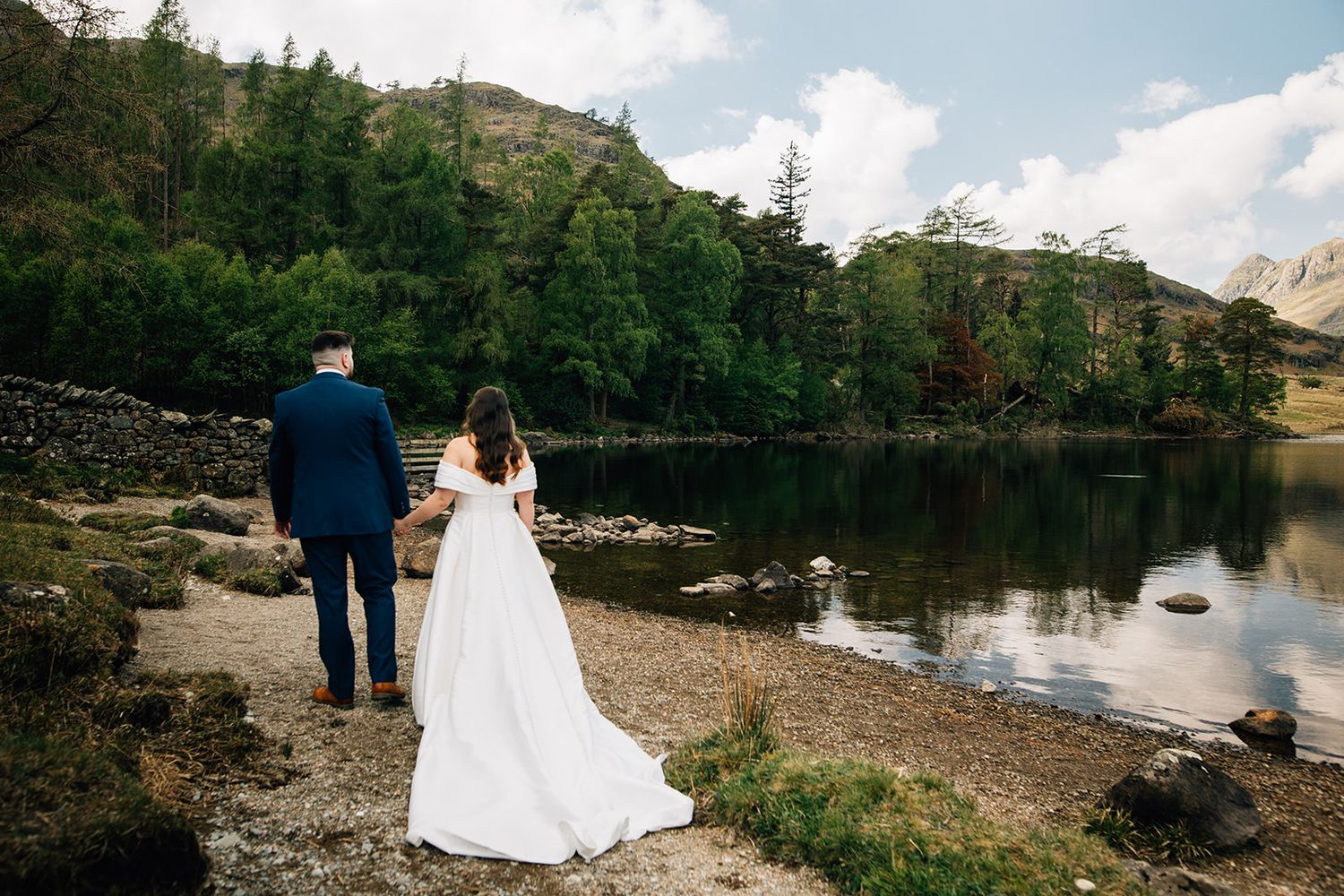 Two people stand at the edge of a serene mountain lake surrounded by evergreen trees on a sunny day.