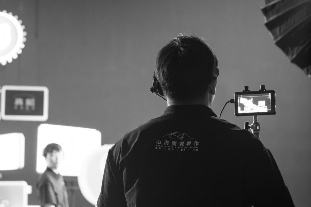 Behind-the-scenes black and white photo showing camera operator filming with lighting equipment in studio setup.