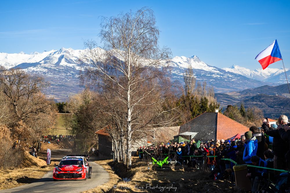 Rally car races along mountain road with snow-capped peaks and spectators watching under Czech flag in background.