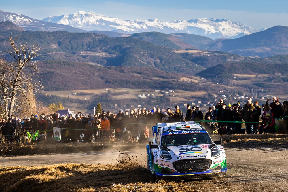 Rally car races through snowy mountain course with snow-capped peaks and rugged terrain in the background.