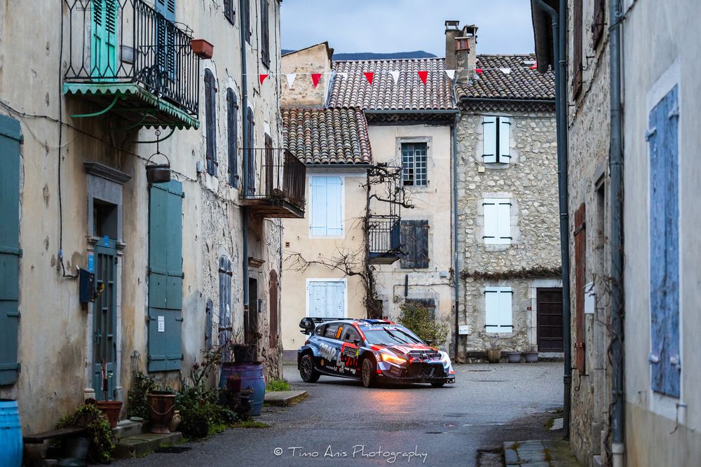 Historic alleyway with stone buildings, red car parked, and traditional architectural details visible.