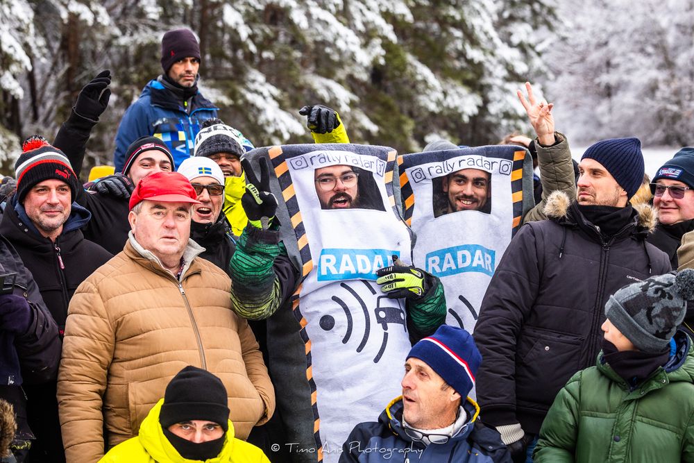 Winter sports spectators gather in snowy conditions wearing warm clothing while holding signs and banners at outdoor event.