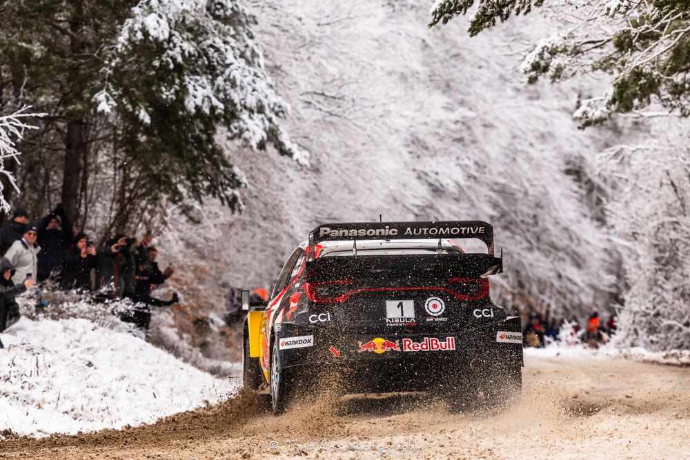 Rally car racing through snowy forest terrain during winter competition event.