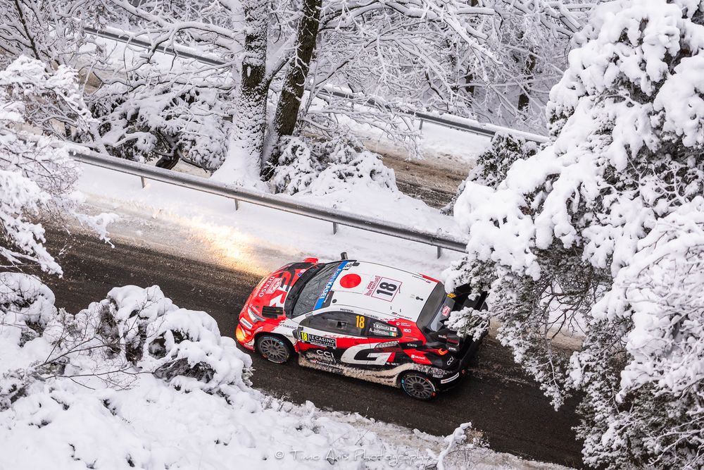 Rally car racing through snowy forest terrain during winter competition on winding mountain road.
