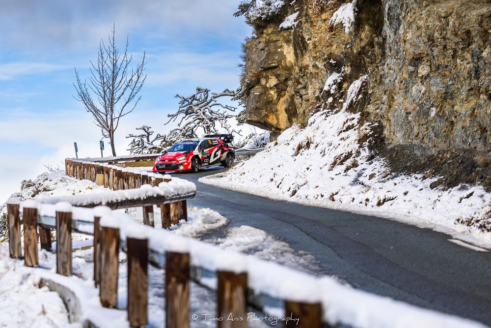 A red sports car drives along a snowy mountain road with rocky cliffs and wooden guardrails on a winter day.