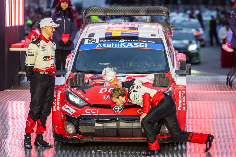 Race cars and pit crews line up in formation on a wet racing track during a motorsport event.