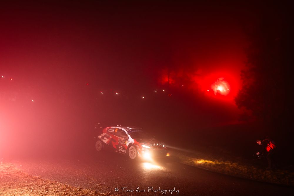 Rally car racing through thick fog at night with red emergency flares illuminating the misty atmosphere.