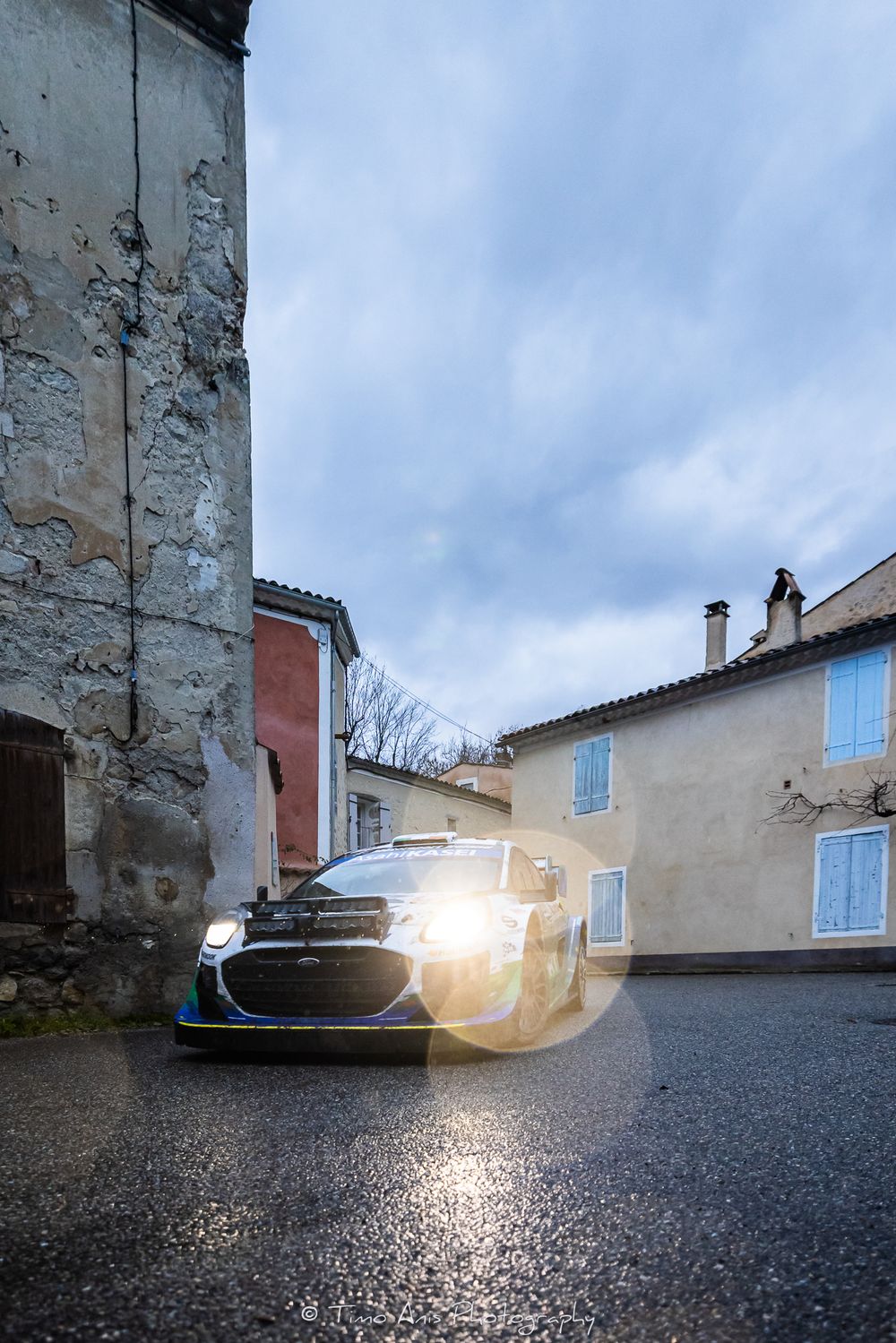 Rally car with bright headlights parked on wet street between old buildings in a French village at dusk.