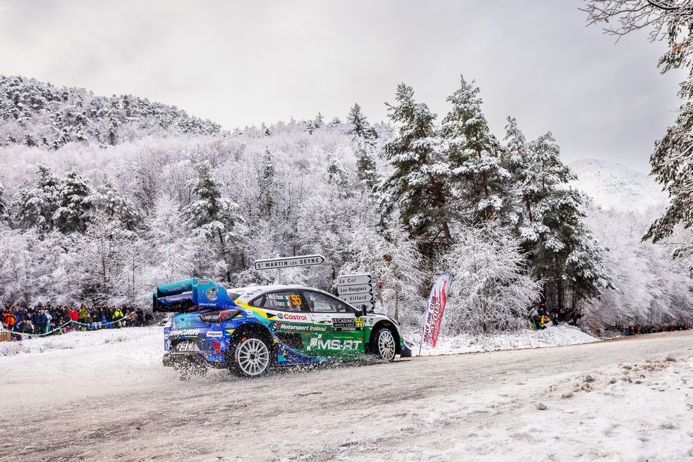 Rally cars race through a snowy winter landscape with frost-covered trees lining the course during a competition.