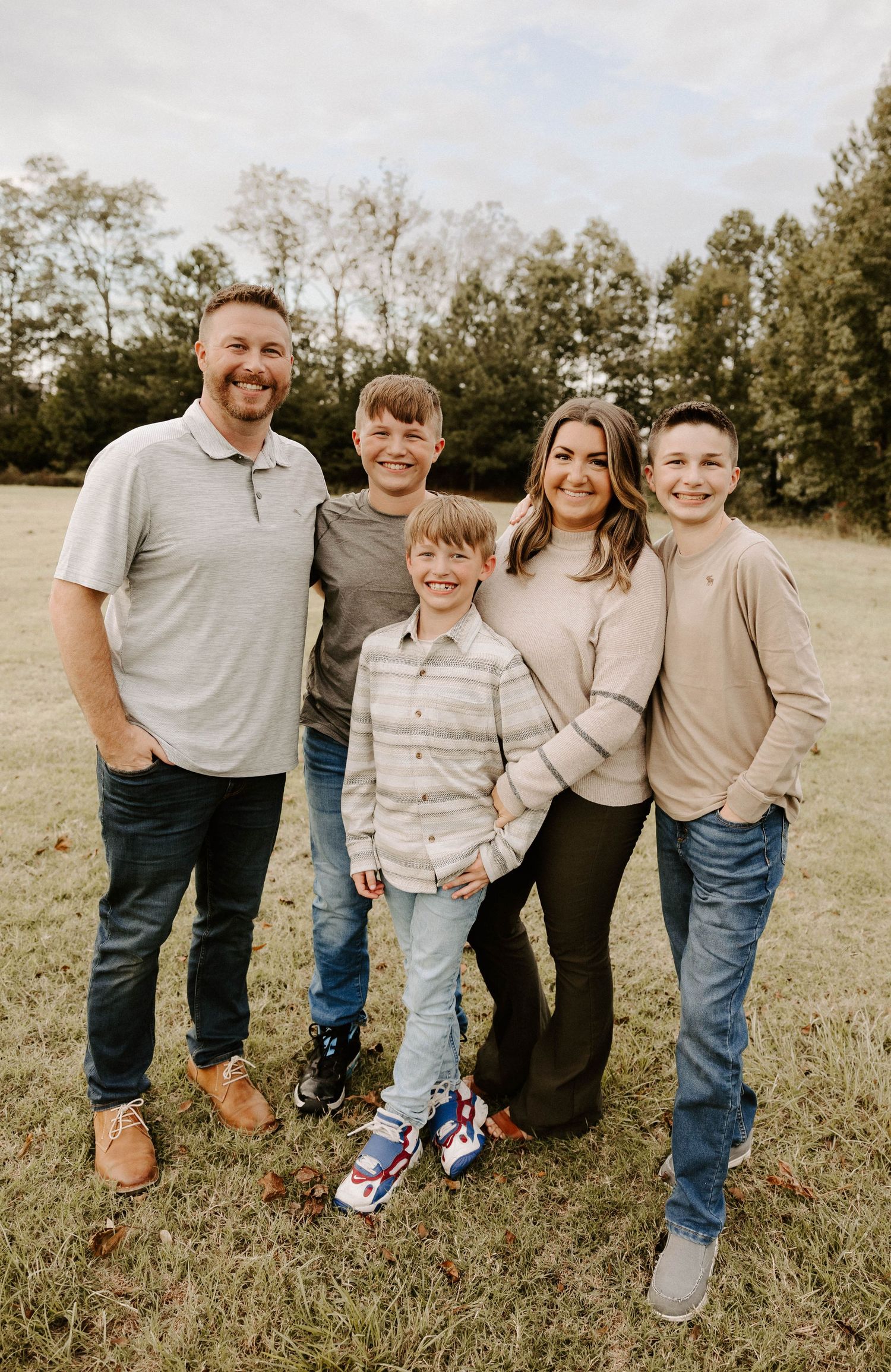 A family group wearing coordinated neutral tones poses together on grassy field during golden hour.