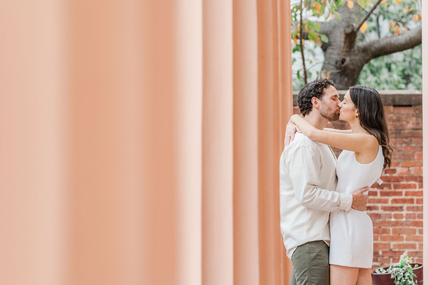 A romantic couple embraces and kisses while standing against a rustic brick wall in an outdoor setting.
